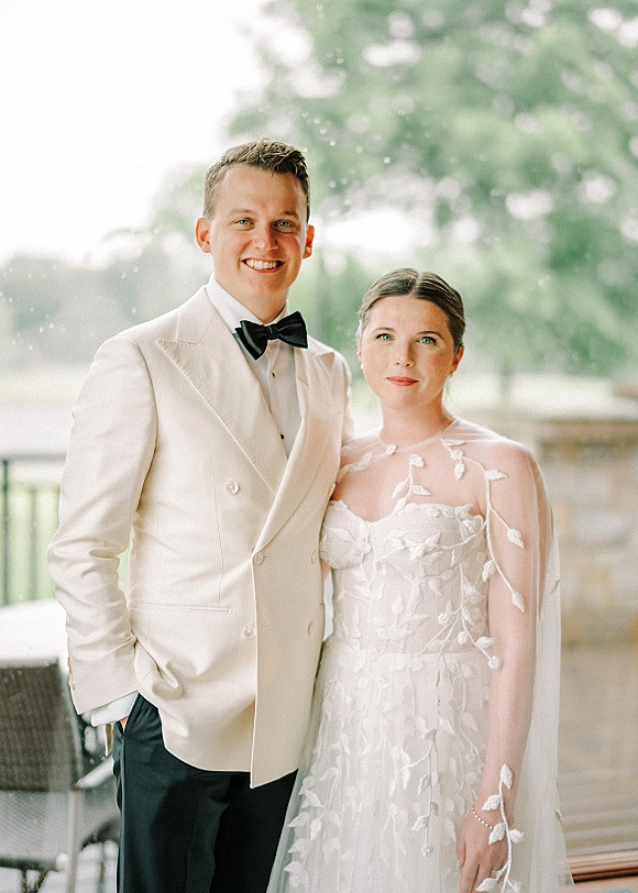 Couple portrait of bride and groom posing by a bright window, her strapless gown with sheer cape and his double-breasted tuxedo.