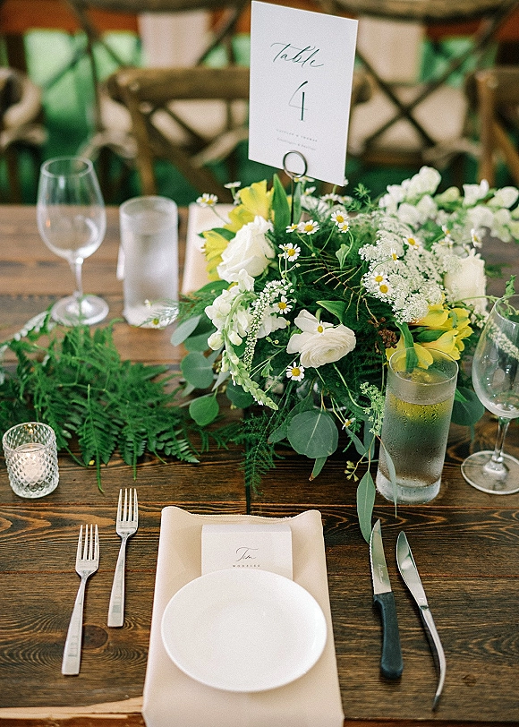 Reception tablescape with wedding table centerpiece and greenery garland on a wood farmhouse table, set with plates, place cards, and votive candle