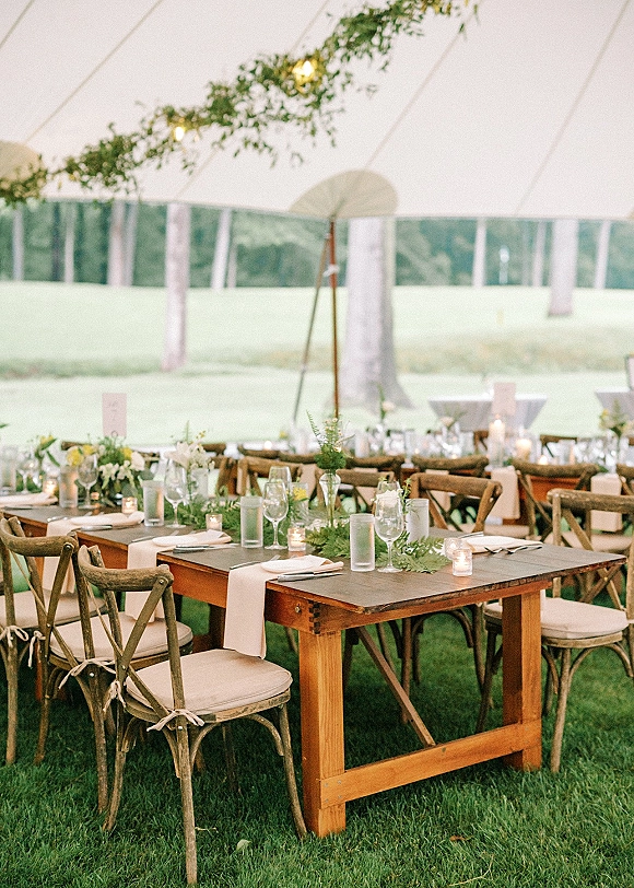 Reception tablescape with greenery garland on wood banquet tables, bud vases, candles and glassware under a clear top tent with string lights