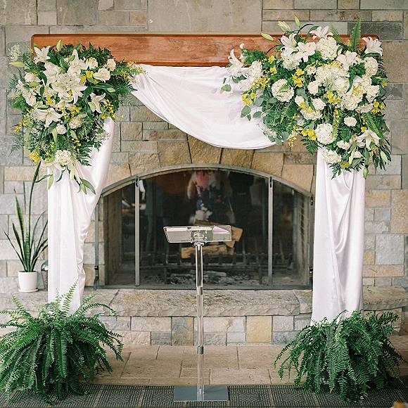 Wedding ceremony arch with white draping fabric and lily-hydrangea florals beside a clear podium, set before a stone fireplace hearth