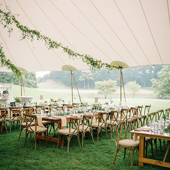 Reception tablescape with outdoor tent reception details, wood farm tables, greenery garland, bud vases, candles, and string lights under a white canopy