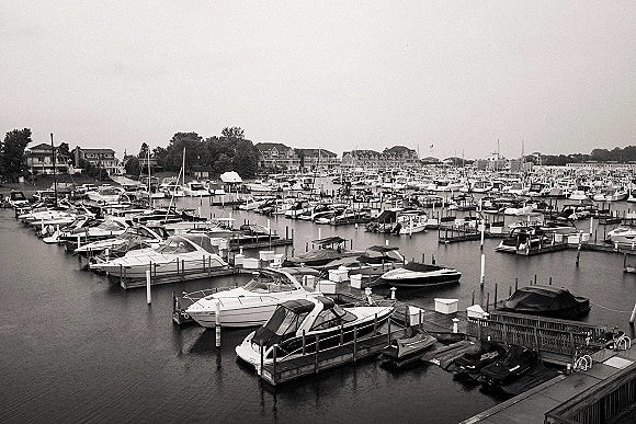 Marina boats moored at docks with sailboat masts rising above calm water, shoreline buildings and trees under an overcast sky