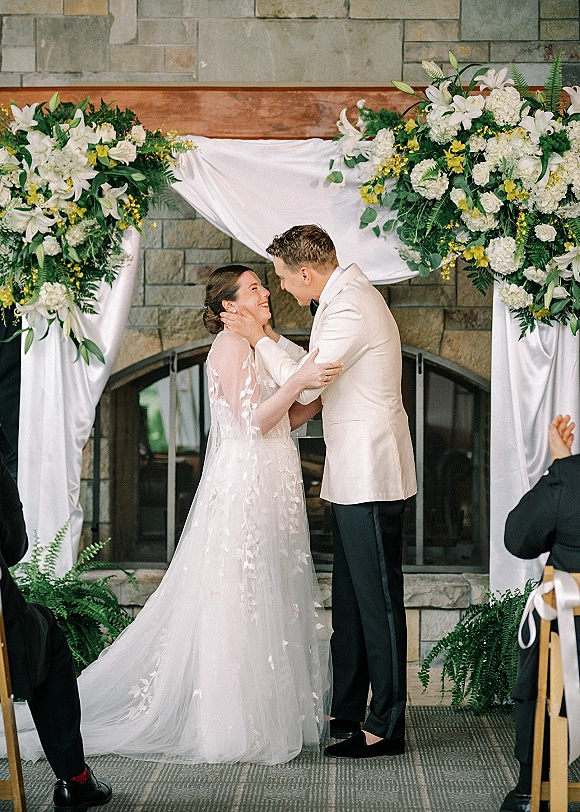 Ceremony moment as bride and groom exchange vows under a wedding ceremony arch with white drapery and lilies, before guests by a stone wall