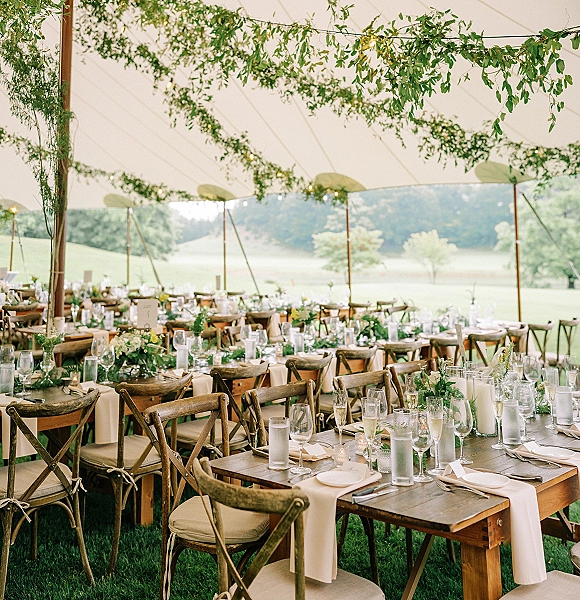 Reception tablescape on farm table wedding reception with greenery garlands, candles, bud vases, and place cards under a sailcloth tent
