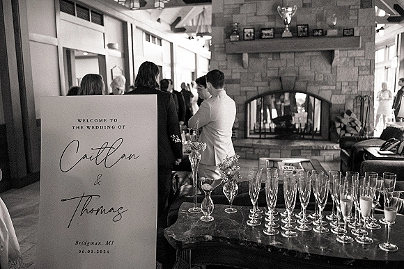 Wedding welcome sign with calligraphy lettering beside champagne flutes and bud vases, set near a stone fireplace at indoor cocktail hour
