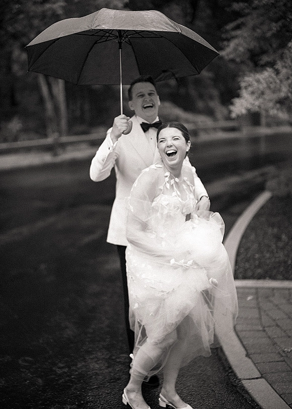 Couple portrait under a wedding umbrella, bride in veil and dress laughing as groom in tuxedo lifts her on a rainy street with trees