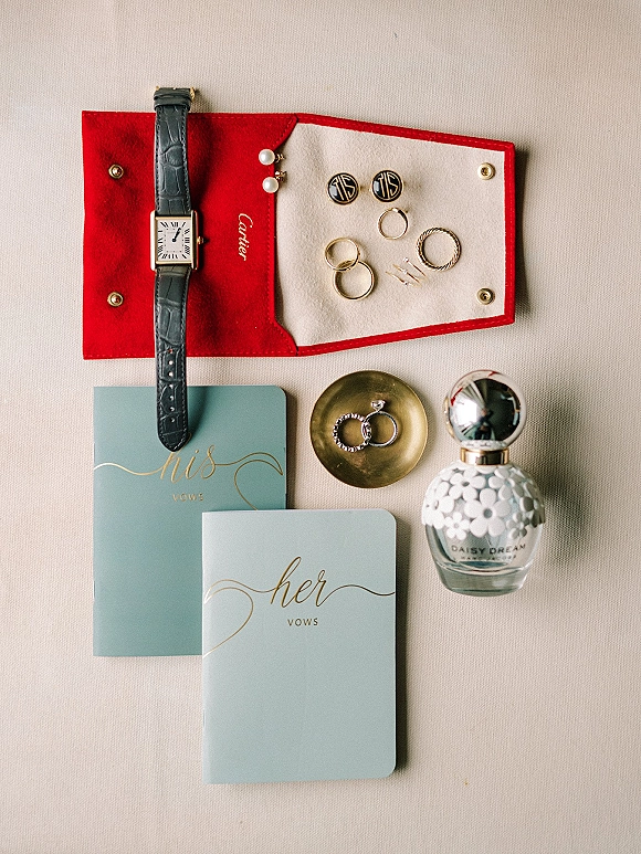 Wedding flat lay of rings, vow books, wristwatch, cufflinks and pearl earrings with a perfume bottle on neutral fabric backdrop