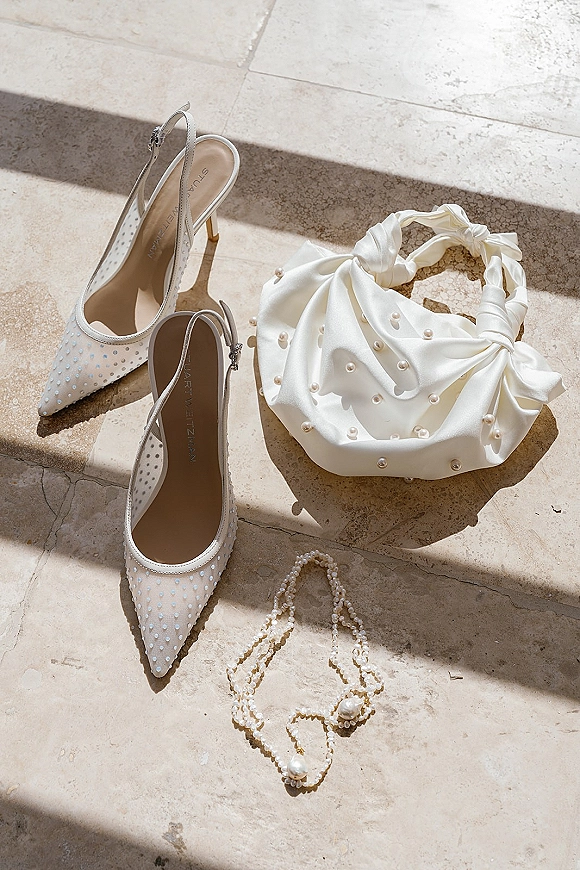Bridal accessories flatlay with wedding shoes flatlay, white pearl slingback heels and satin clutch on stone floor in sunlit shadows