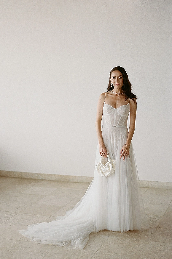Bridal portrait of a bride in a simple wedding dress with corset bodice, tulle skirt and pearl belt, holding a satin clutch by a white wall
