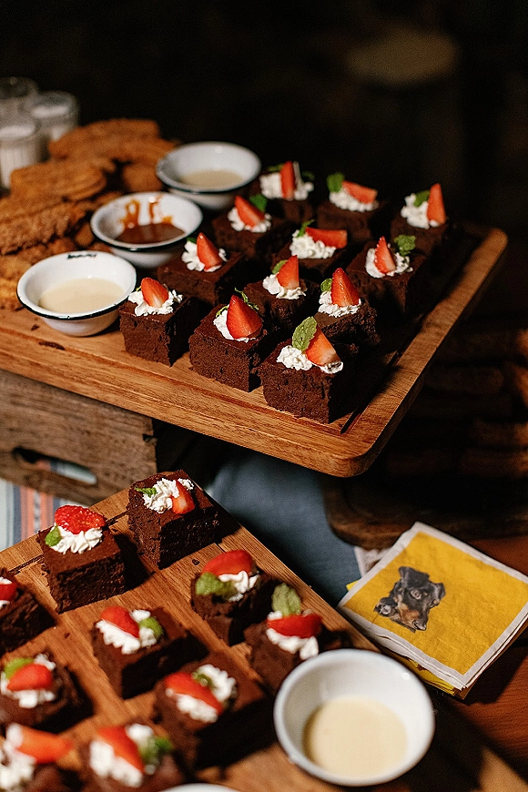 Wedding dessert table with brownie dessert bar bites, strawberries and whipped cream on rustic wooden boards against a dark backdrop
