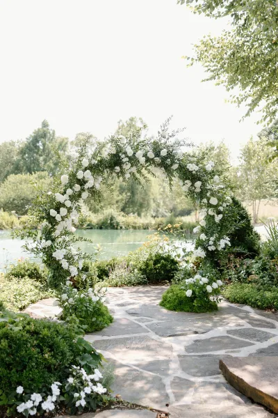 Wedding ceremony arch with circle wedding arch design, white roses and greenery on moss base, set on stone patio by a lake and trees
