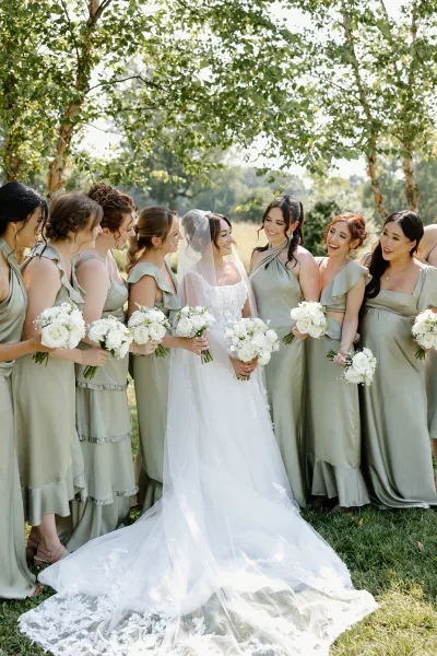 Bride with bridesmaids in sage dresses laughing together, holding white rose bouquets on a sunlit lawn with trees in the background