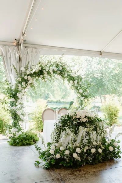 Wedding sweetheart table with sweetheart table backdrop of white roses and greenery garland under string lights in a draped white tent on lawn