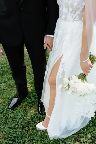 Wedding couple portrait of bride and groom holding hands, showcasing lace wedding dress with bouquet and black tuxedo on lawn