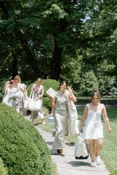 Bridal party candid as bridesmaids walking together with tote bags along a stone path through garden hedges, bride in slip dress behind