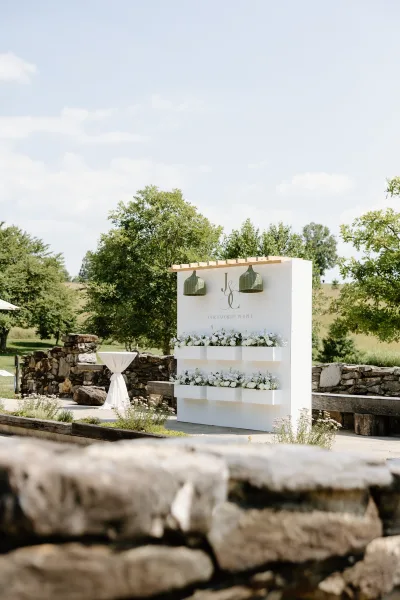 Wedding escort display with an escort card wall and built-in white floral boxes under a wooden canopy with pendant lights by a stone wall lawn
