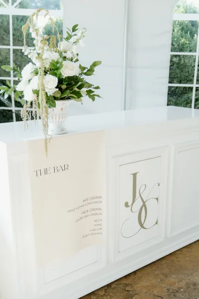 Wedding bar setup with a wedding bar sign and white rose floral arrangement in an urn vase atop a white counter inside a tented reception