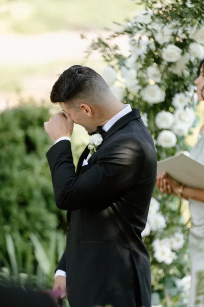 Ceremony moment as groom cries at altar, wiping tears in a black tuxedo beneath a white rose and greenery floral arch outdoors