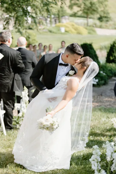 Wedding kiss portrait of newlyweds as the groom dips the bride, her veil flowing beside white aisle florals in a garden ceremony setting
