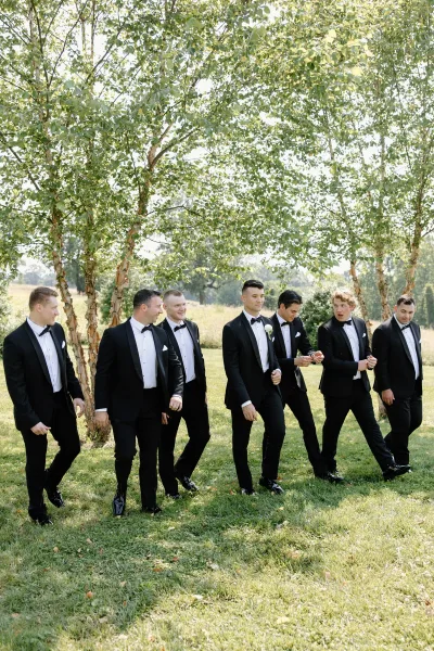 Groomsmen portrait with men in black tuxedos and bow ties, standing on a grassy lawn by birch trees under daylight sky