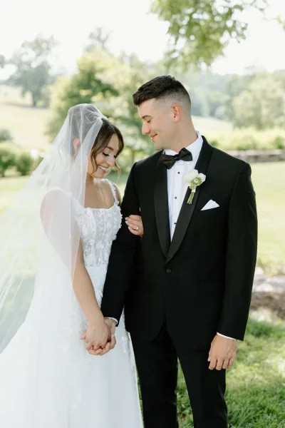 Couple portrait of bride and groom holding hands, looking down, with a flowing veil on a green lawn beside trees and a stone wall