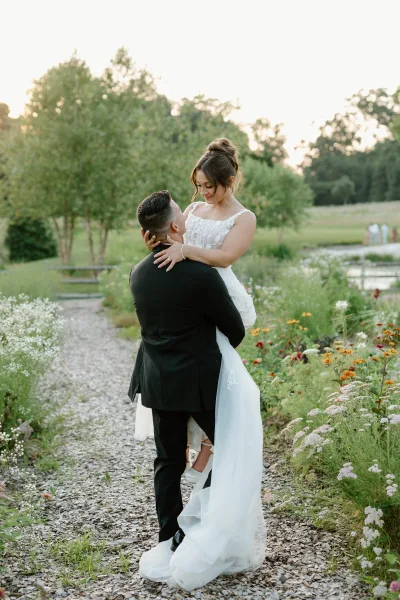 Couple portrait with groom lifting bride in his arms, her lace wedding dress and long veil flowing along a garden path by wildflowers and pond