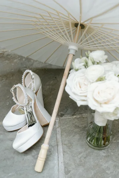 Bridal accessories flat lay with white platform heels and pearl ankle straps beside a white parasol and rose bouquet on a stone patio