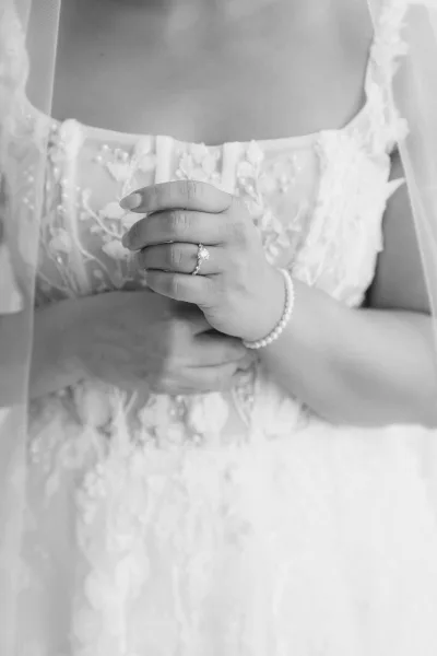 Bridal ring close-up of an oval diamond engagement ring on the bride’s left hand, with lace wedding dress bodice detail and pearl bracelet