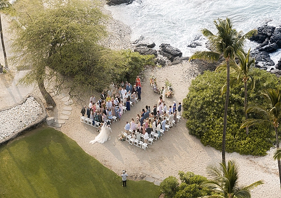 Wedding ceremony on beach with bride walking down the aisle toward groom, white folding chairs in a circle beside ocean waves and palms