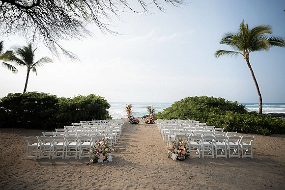 Beach ceremony setup with white folding chairs lining a sand aisle, pastel floral markers and ocean waves, palms and sky beyond