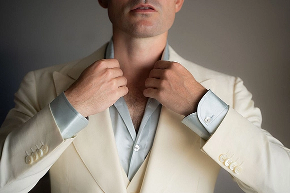 Groom getting ready, adjusting collar on a cream suit jacket with cufflinks and buttons against a neutral wall backdrop