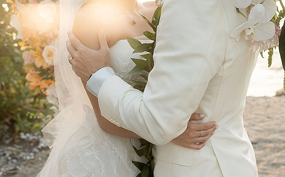Wedding couple portrait of bride and groom embrace, her bridal veil and lace bodice visible on a sunlit beach with ocean behind