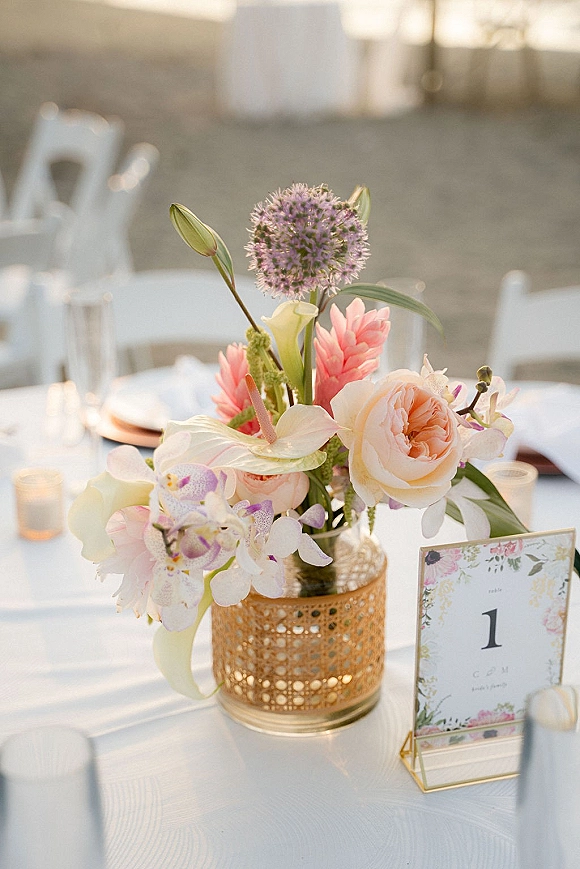 Wedding centerpiece with pastel roses, orchids and calla lilies in a gold vase, surrounded by tealights and a table number on a white cloth outdoors