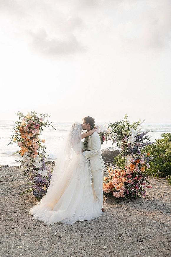 Wedding kiss portrait of bride and groom kissing under a floral arch, her long veil flowing, with ocean waves and cloudy sky behind