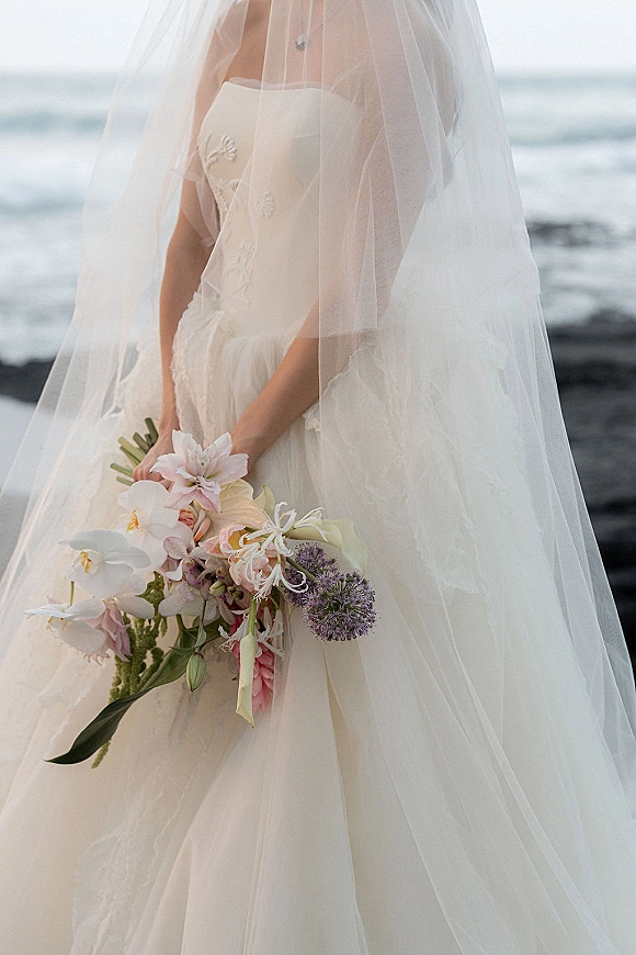 Bridal portrait of a bride holding bouquet with a veil over her face, strapless dress, and orchid lilies by the ocean shoreline