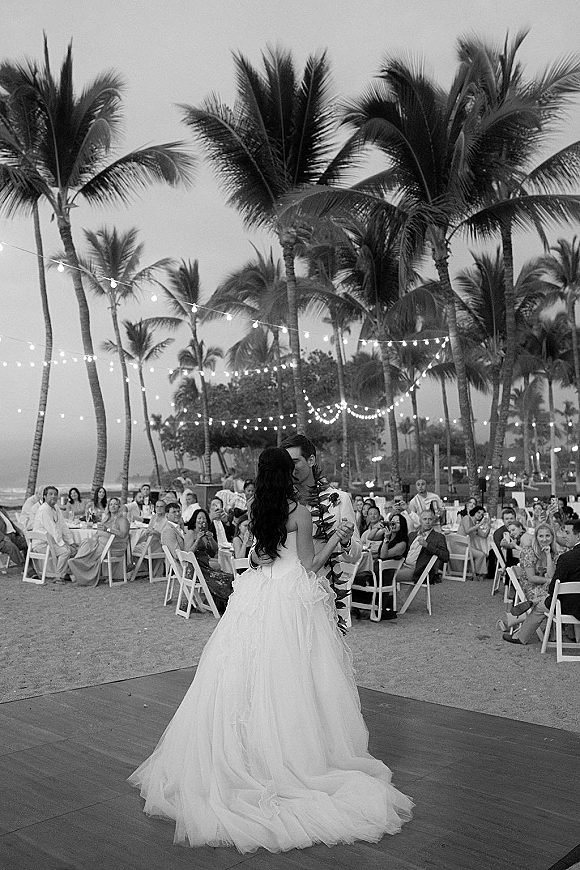 First dance of bride in a wedding gown and groom in suit with floral lei under string lights on a beach dance floor by palm trees