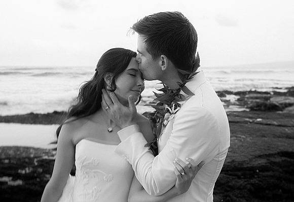 Wedding couple portrait of bride and groom embrace as he kisses her forehead, lei accent and rings visible on a rocky ocean shoreline