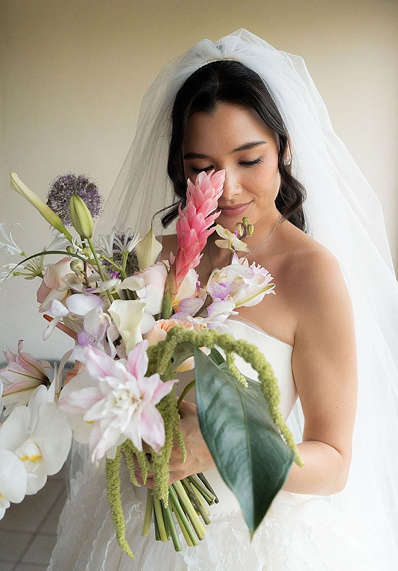 Bridal portrait of a bride holding bouquet with veil, strapless dress and tropical orchids and calla lilies against a neutral wall