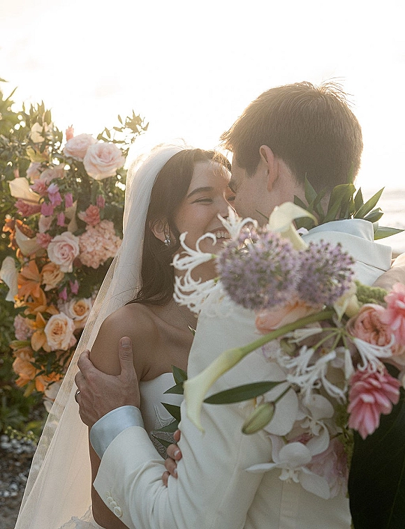 Wedding kiss as bride and groom embrace under a floral arch, her veil flowing beside an ocean coastline at sunset