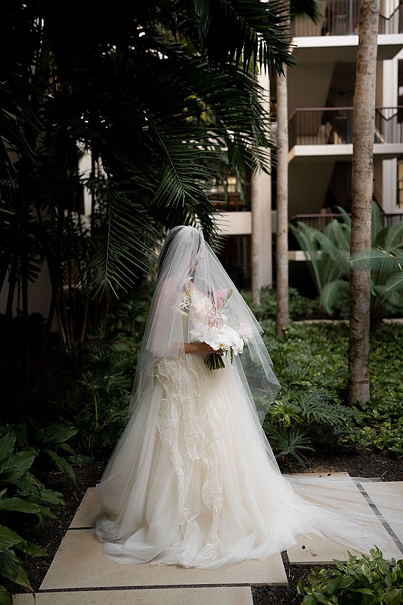 Bridal portrait of a bride holding bouquet with a veil over her face, lace strapless gown, and palm-lined garden walkway backdrop