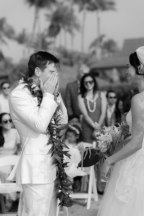First look moment as groom crying covers his face in a white suit while bride in veil holds bouquet at palm tree ceremony