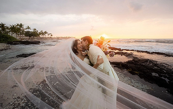 Wedding couple portrait at sunset beach, bride in strapless gown holding bouquet with veil blowing as groom hugs her by ocean waves