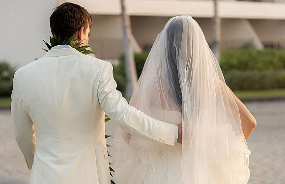 Couple portrait of bride and groom from behind, groom’s arm around her as they walk away on an outdoor lawn by a building, long veil flowing