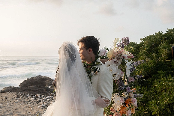 Wedding couple portrait of bride and groom embracing under a floral arch, her veil and bouquet flowing by the rocky ocean shoreline