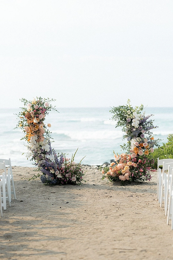 Beach ceremony setup with oceanfront wedding ceremony aisle, white folding chairs and floral pillars in pastel blooms on sand by the shoreline