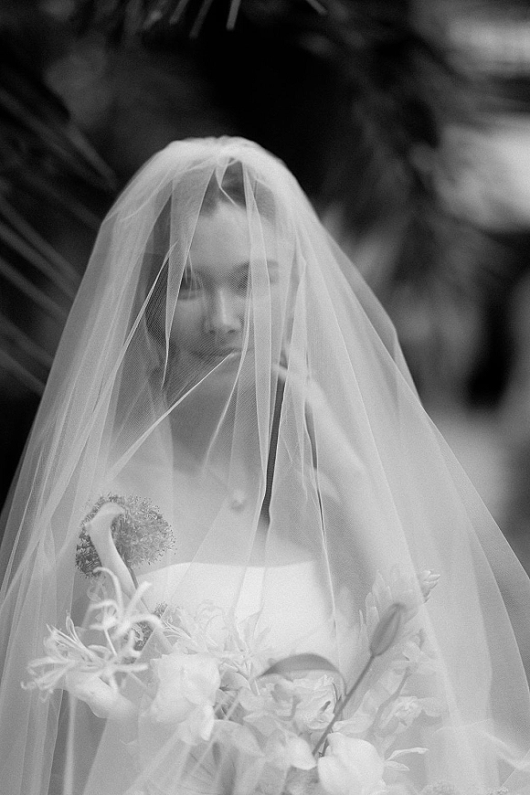 Bridal portrait of a bride with veil draped over her face, holding a bouquet in a strapless dress against lush outdoor greenery