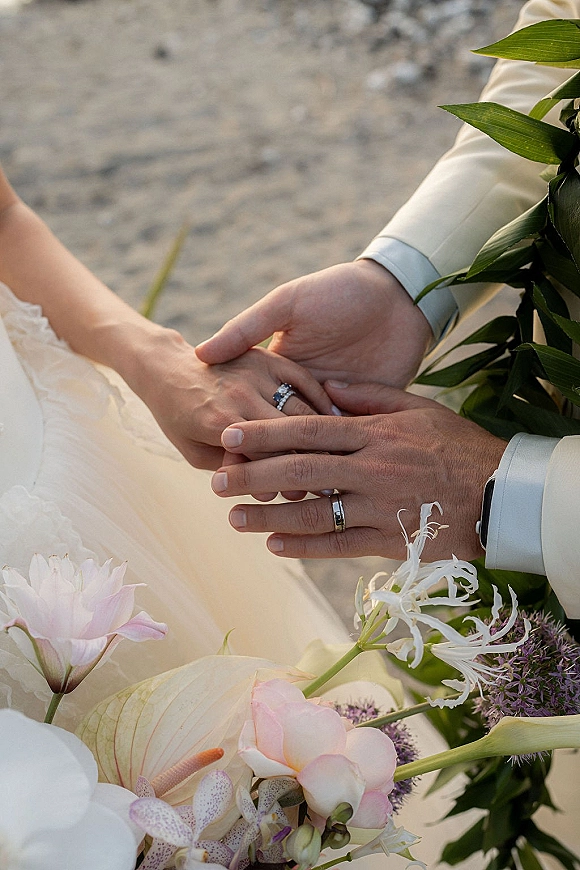 Wedding rings close-up with engagement ring sparkle as bride and groom hands hold a ring stack beside a blush bouquet on stone ground