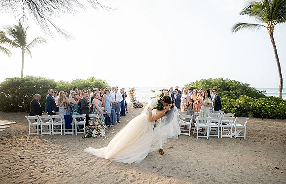 Wedding kiss as groom dips bride in strapless dress and veil, bouquet in hand, on sandy aisle with chairs, palms and ocean beyond