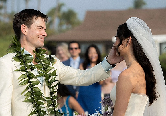 Ceremony moment as groom touches bride’s face during vows, her veil and strapless dress visible, guests blurred with palm trees behind