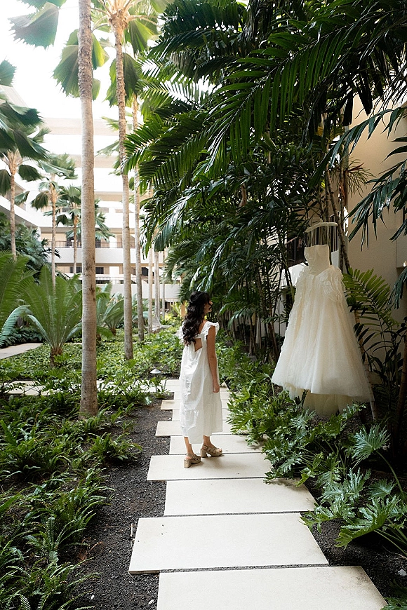 Wedding dress on hanger, a strapless tulle bridal gown hanging in a tropical hotel courtyard with palm trees and stone walkway